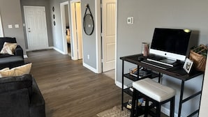 A living room with a dark wooden floor, a black sofa, a white door, and a desk with a computer. - Cozy Corner <7 miles from Toyota Field and Redstone Arsenal - family-friendly! (Madison)