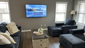 A living room with a flat-screen TV mounted on the wall, a coffee table with a vase, and two dark-colored sofas. - Cozy Corner <7 miles from Toyota Field and Redstone Arsenal - family-friendly! (Madison)