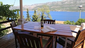 Une table en bois, deux chaises, une bouteille et deux verres sont disposés sur une terrasse qui offre une vue sur un plan d’eau et des montagnes.