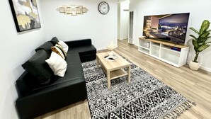 A modern living room with a black sectional sofa, a wooden coffee table, a patterned rug, a flat-screen TV, and a wall clock. -  Charming House in Old Town Fullerton near Disney & Knott’s!! (Fullerton)