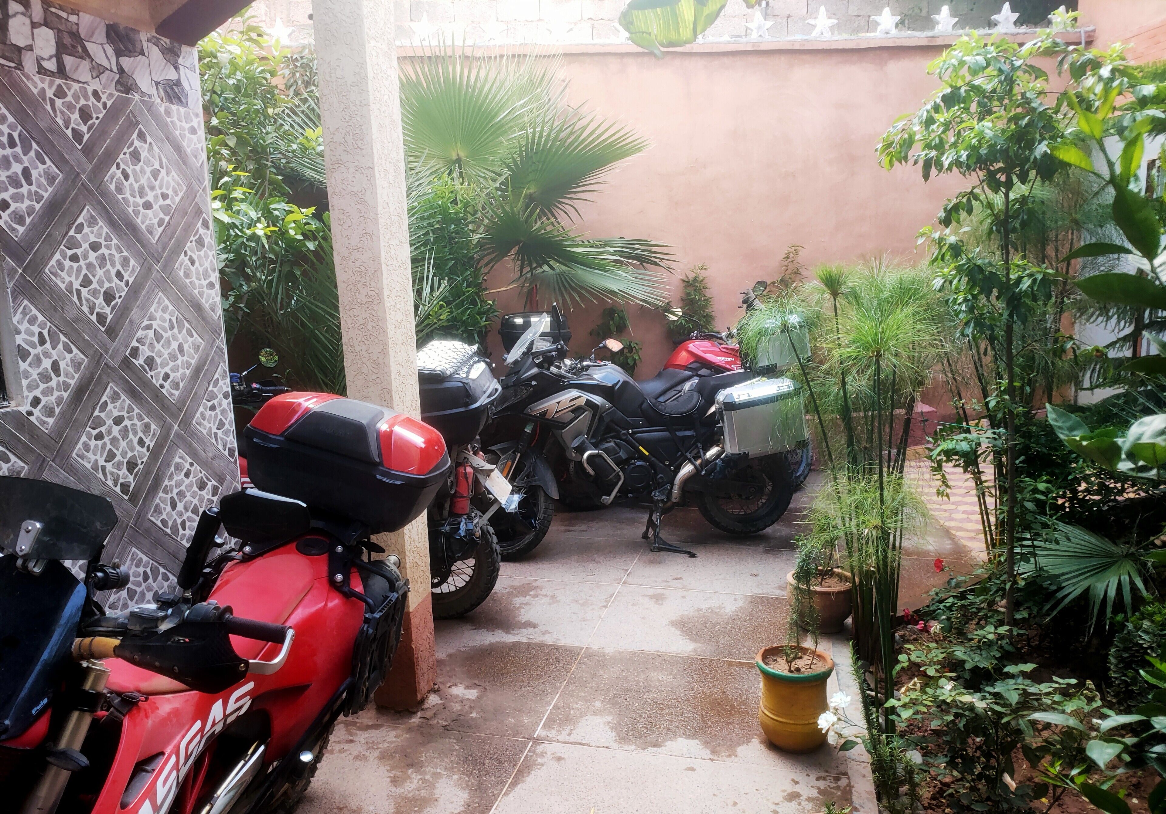 A courtyard with motorcycles parked along the walls, surrounded by potted plants and a tiled floor.