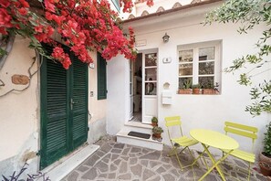 Un bâtiment blanc avec des volets verts, une entrée pavée de pierres et une table jaune avec des chaises.
