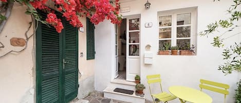 A white building with green shutters, a stone-paved entrance, and a yellow table with chairs.