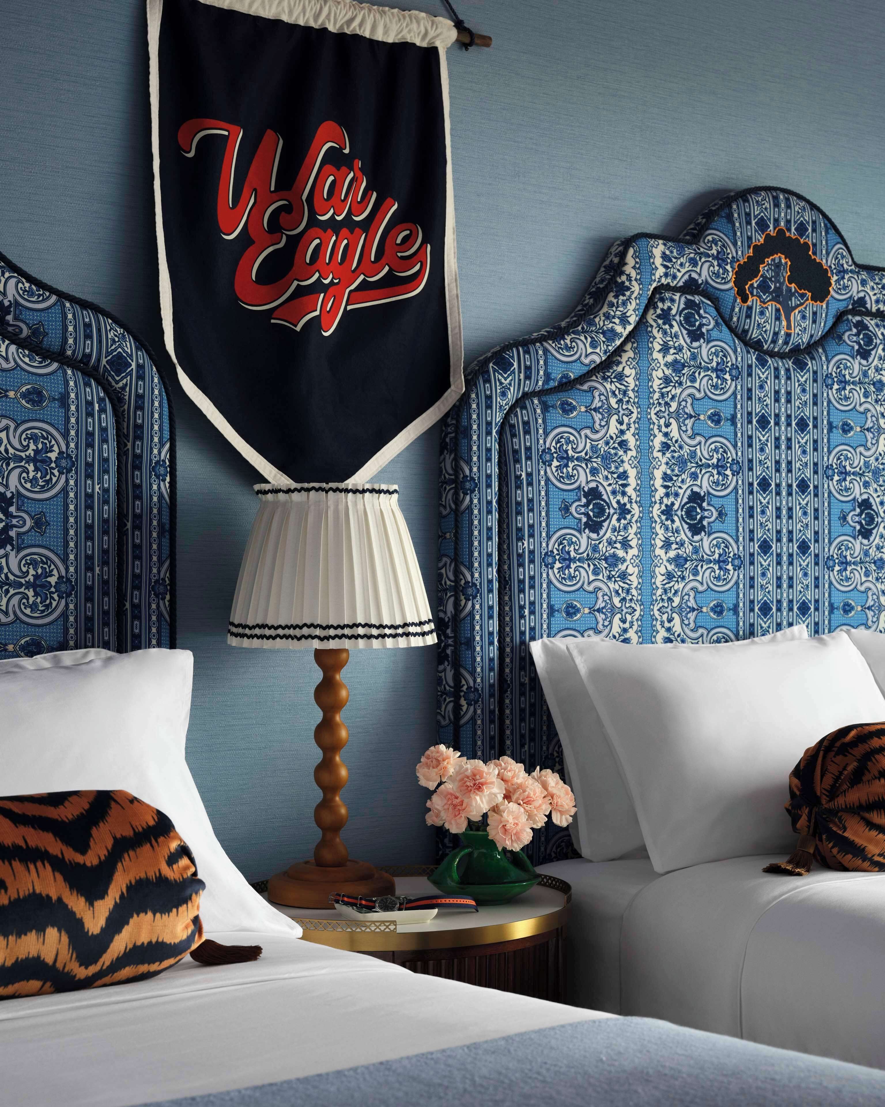 Una habitación de hotel con una cama, una mesita de noche, una lámpara, un florero con flores y un tapiz con el texto "Viva Eagle".