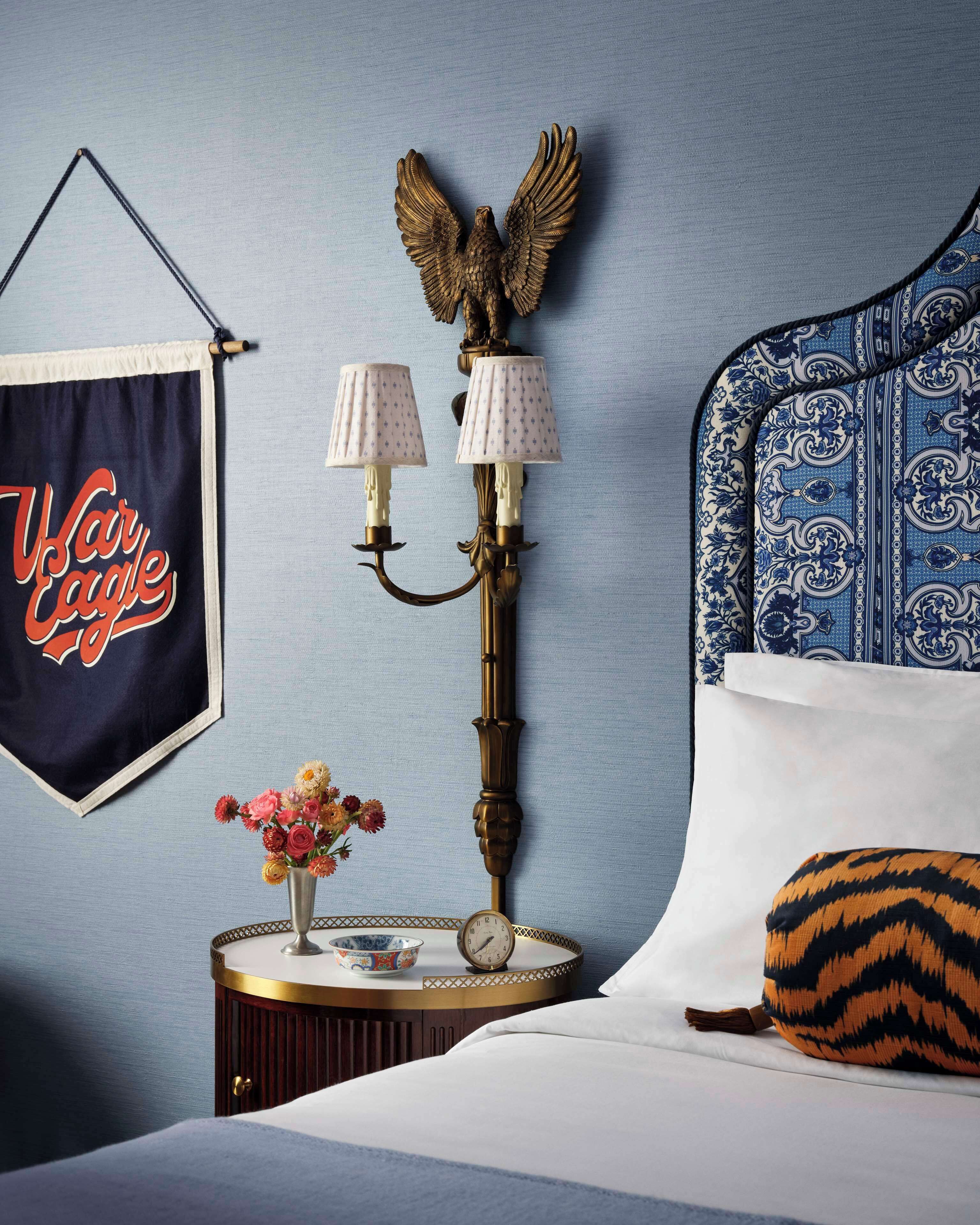 Een slaapkamer met een bed, nachtkastje met een klok, een wandlamp, een lijst en een vaas met bloemen.