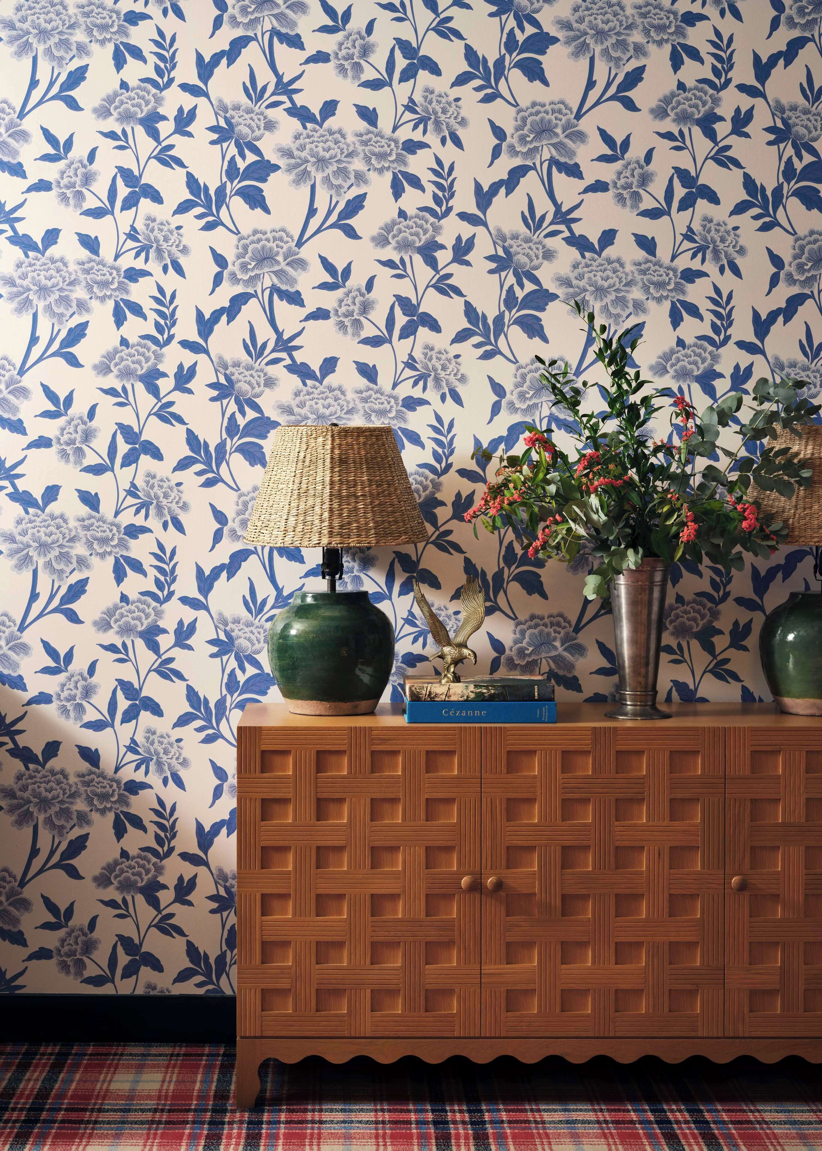 Un meuble en bois, avec un papier peint à motifs floraux en arrière-plan, une lampe en osier, un vase vert et un livre intitulé « Cézanne ».