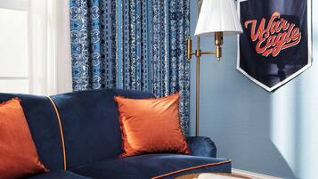 Una habitación con un sofá azul oscuro, cojines naranjas, una mesita redonda de madera con un libro, una lámpara y un cuadro con la imagen de 'War Eagle' en la pared.