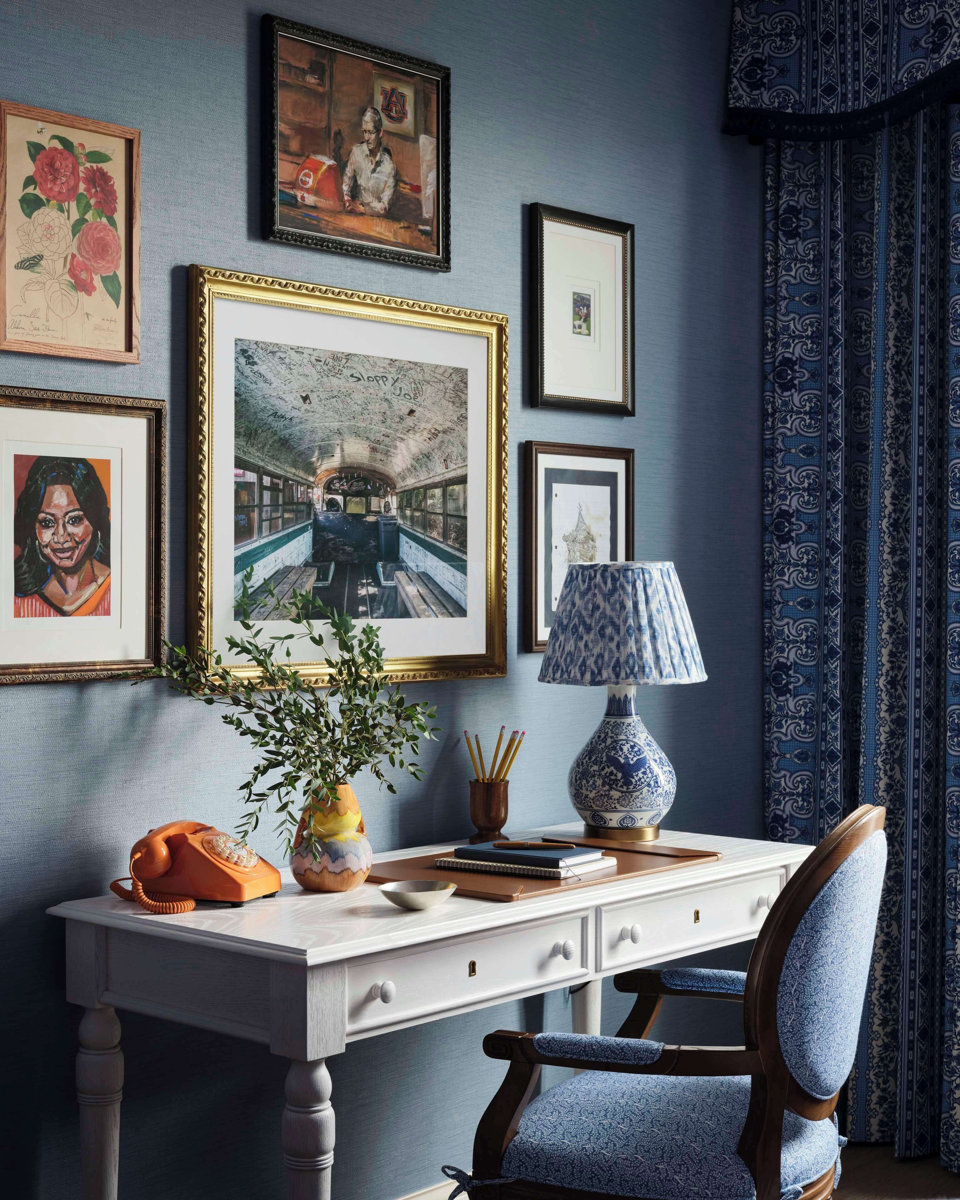 Um quarto com escrivaninha, cadeira, quadros e abajur.