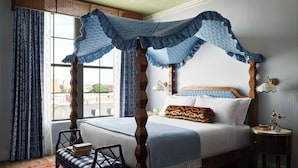 A four-poster canopy bed with a view of the city through large windows, a bedside table with a vase of flowers, and a patterned rug. - Graduate by Hilton Auburn AL (Auburn)