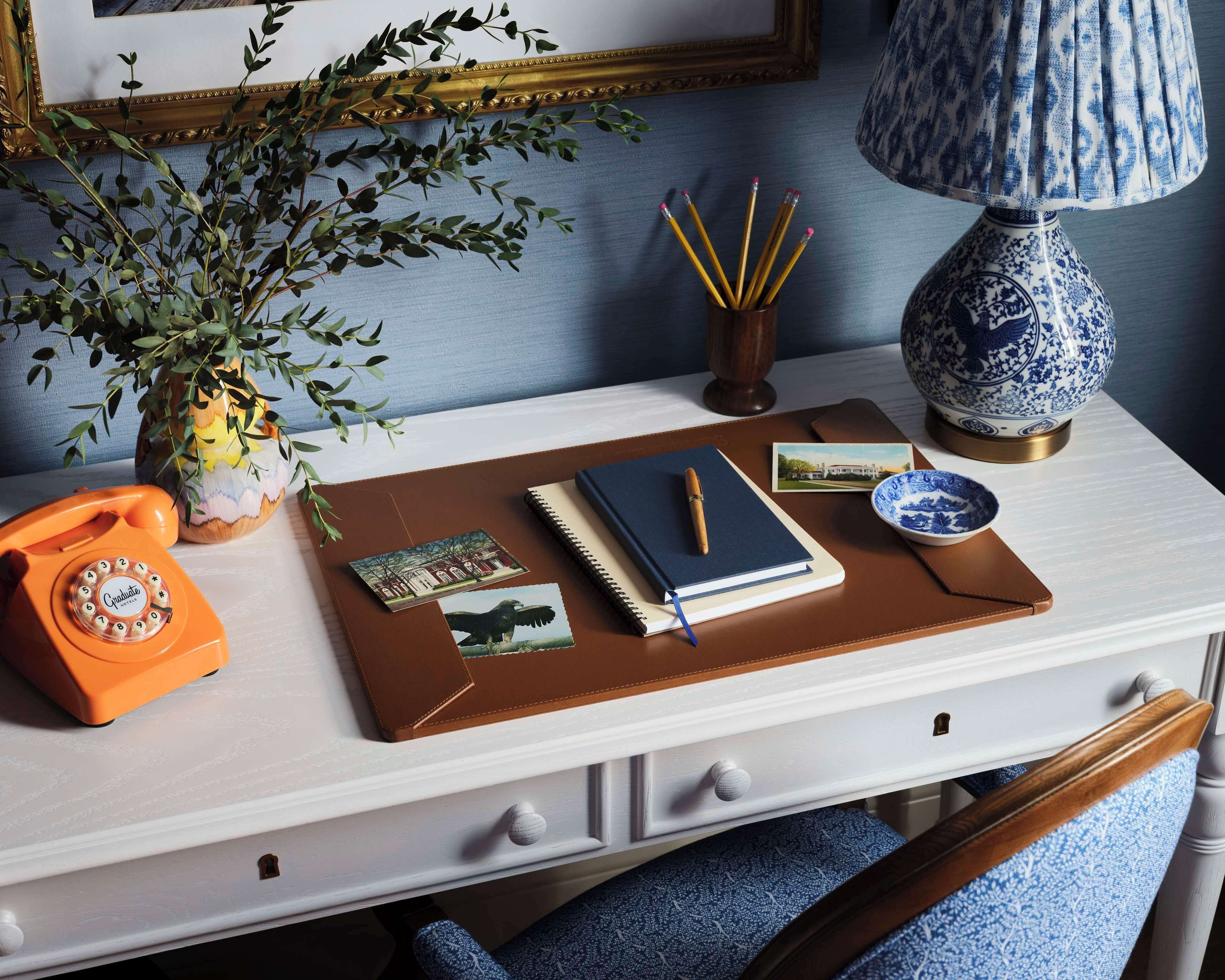 A desk with a rotary phone, a notebook, a pen, a lamp, and a vase with a plant.