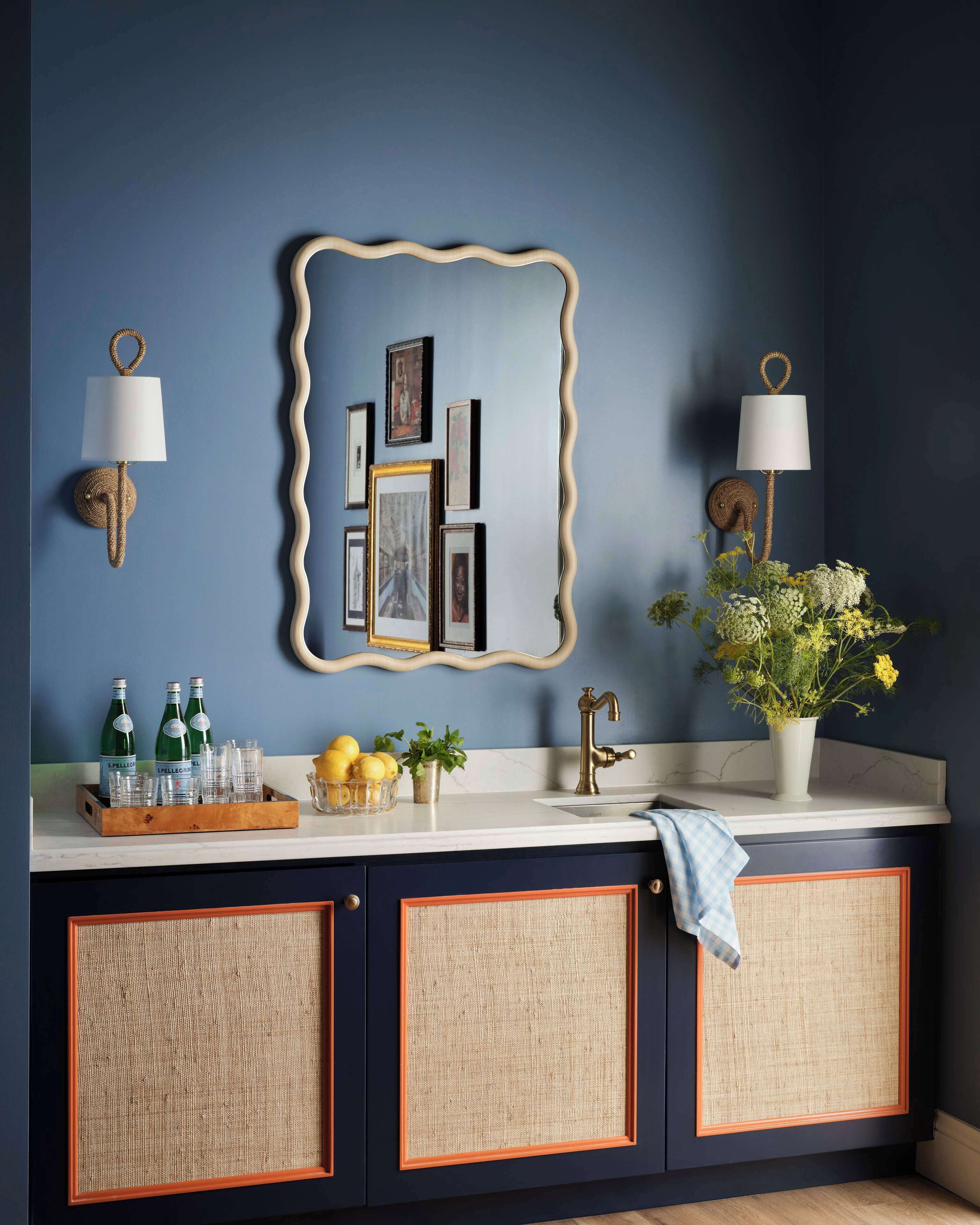 Une cuisine avec un mur bleu, un miroir décoratif, des lampes fixées au mur, un évier avec un robinet et un saladier rempli de citrons et agrémenté d’une plante.