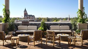 A rooftop terrace with a view of a historic building, furnished with wicker chairs and a sofa, and a wooden deck. - Graduate by Hilton Auburn AL (Auburn)