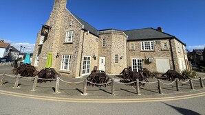 Un bâtiment en pierre doté d’une cheminée imposante, de nombreuses fenêtres et d’un espace pavé aménagé avec des chaises et des tables en bois.