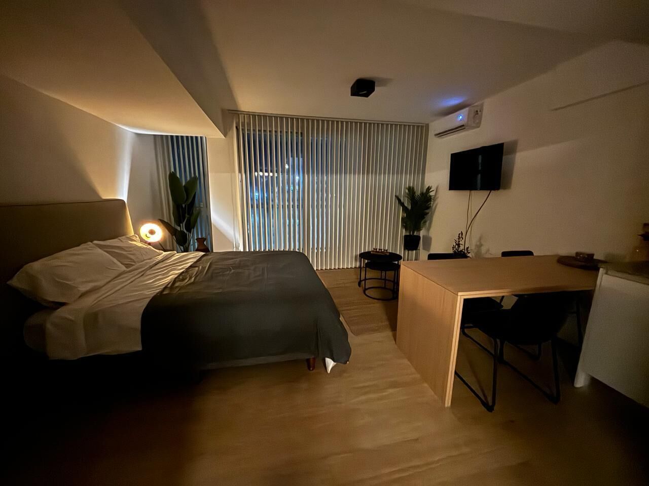 A bedroom with a bed, bedside table, desk, and chair. There is a window with blinds, a plant, and a TV mounted on the wall.