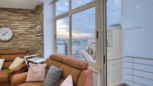 A modern living room with a large window, a clock, and a city view. - Roof terrace, Seaview 3 bedroom penthouse GOSLM-11 (Sliema)