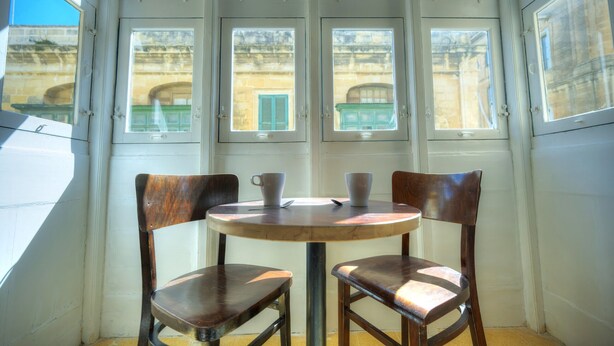 Una habitación de esquina con una mesa redonda y dos sillas, ventanas amplias con vistas a edificios y una puerta.