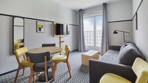 Ein modernes Hotelzimmer mit einem runden Esstisch, zwei gelben Stühlen, einem grauen Sofa, einem an der Wand montierten Fernseher und einem Fenster mit Vorhängen.