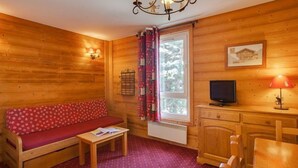 A cozy cabin room with a wooden interior, a sofa, a small table, a TV, and a window with curtains.