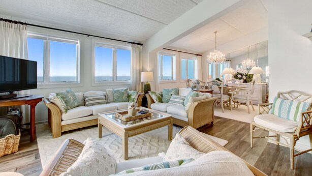 A spacious living room with a large flat-screen TV, a sofa set, a coffee table, wicker chairs, and a dining area with a chandelier. - 3 Bedroom/3Bath Oceanfront PentHouse in Shipwatch (Fernandina Beach)