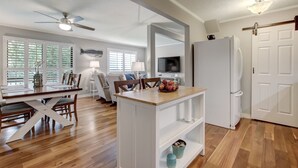Ein geräumiger Wohnbereich mit einem Holzesstisch, einer weißen Kücheninsel mit einer Lampe, einem Deckenventilator und einem weißen Kühlschrank.