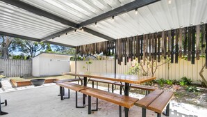 Een overdekte terras met een picknicktafel en bankjes, omgeven door een houten hek en beplanting.