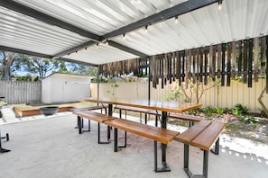 Un patio techado al aire libre con una mesa de picnic y bancos, rodeado por una cerca de madera y plantas.
