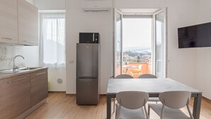 Uma cozinha moderna com geladeira, mesa de jantar com cadeiras, TV fixada na parede e janela com vista para edifícios.