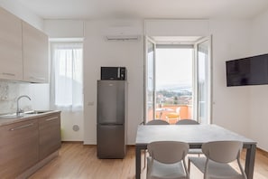 A modern kitchen with a refrigerator, a dining table with chairs, a TV mounted on the wall, and a window with a view of buildings.