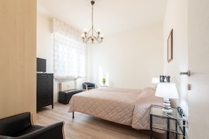 Camera da letto con un letto, una scrivania, una sedia, una cassettiera, una TV, un lampadario e una finestra con tende.