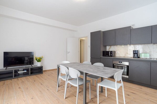 A modern kitchen with dark cabinets, a built-in oven, and a microwave. There is a dining table with four chairs and a flat-screen TV on a stand.