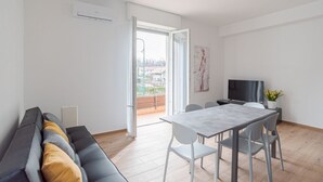 A modern living room with a sofa, a dining table with chairs, a TV, and a balcony with a view.