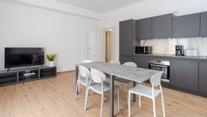 A modern kitchen with dark cabinets, a built-in oven, and a microwave. There is a dining table with four chairs and a flat-screen TV on a stand.