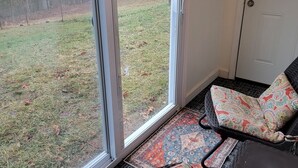 A room with a sliding glass door leading to a grassy yard, a black wicker chair with colorful cushions, and a patterned rug on the floor. - Lake Cabin, Fishing Access, swimming, walk to lake, boat parking, fenced yard!  (Nancy)