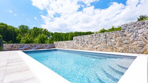Una piscina rectangular con paredes de piedra y solárium con baldosas, rodeada de árboles.