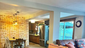 Ein Wohnzimmer mit braunem Sofa, ein Essbereich mit Holztisch und -stühlen sowie eine Küche mit Kühlschrank und Schränken.