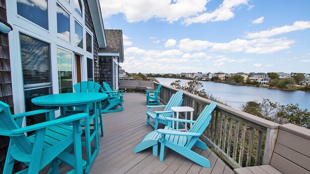Una terraza de madera con sillas Adirondack turquesa y una pequeña mesa, con vista a un cuerpo de agua y casas a lo lejos.