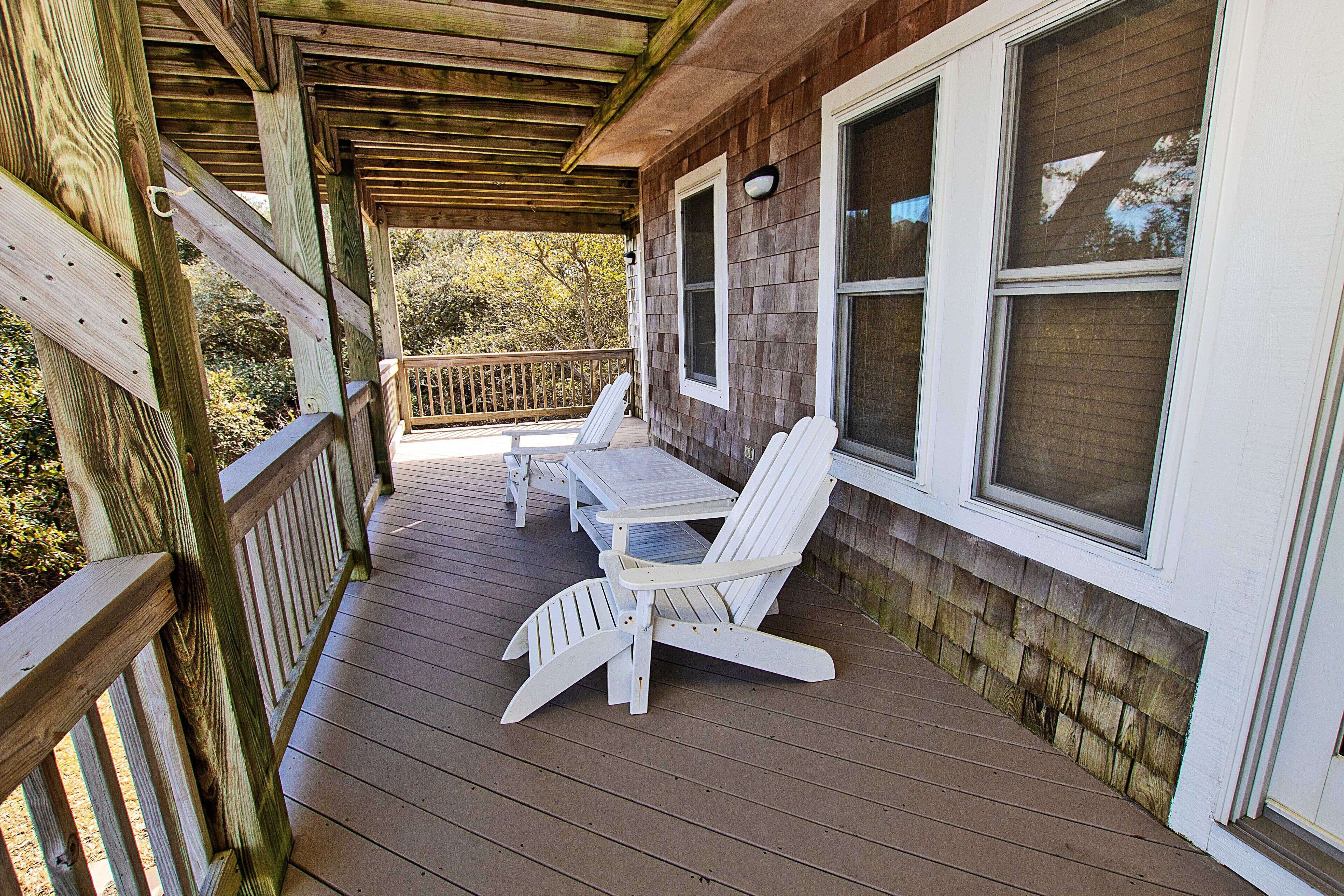 A wooden deck with two Adirondack chairs and a table, overlooking a wooded area.