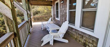 A wooden deck with two Adirondack chairs and a table, overlooking a wooded area.