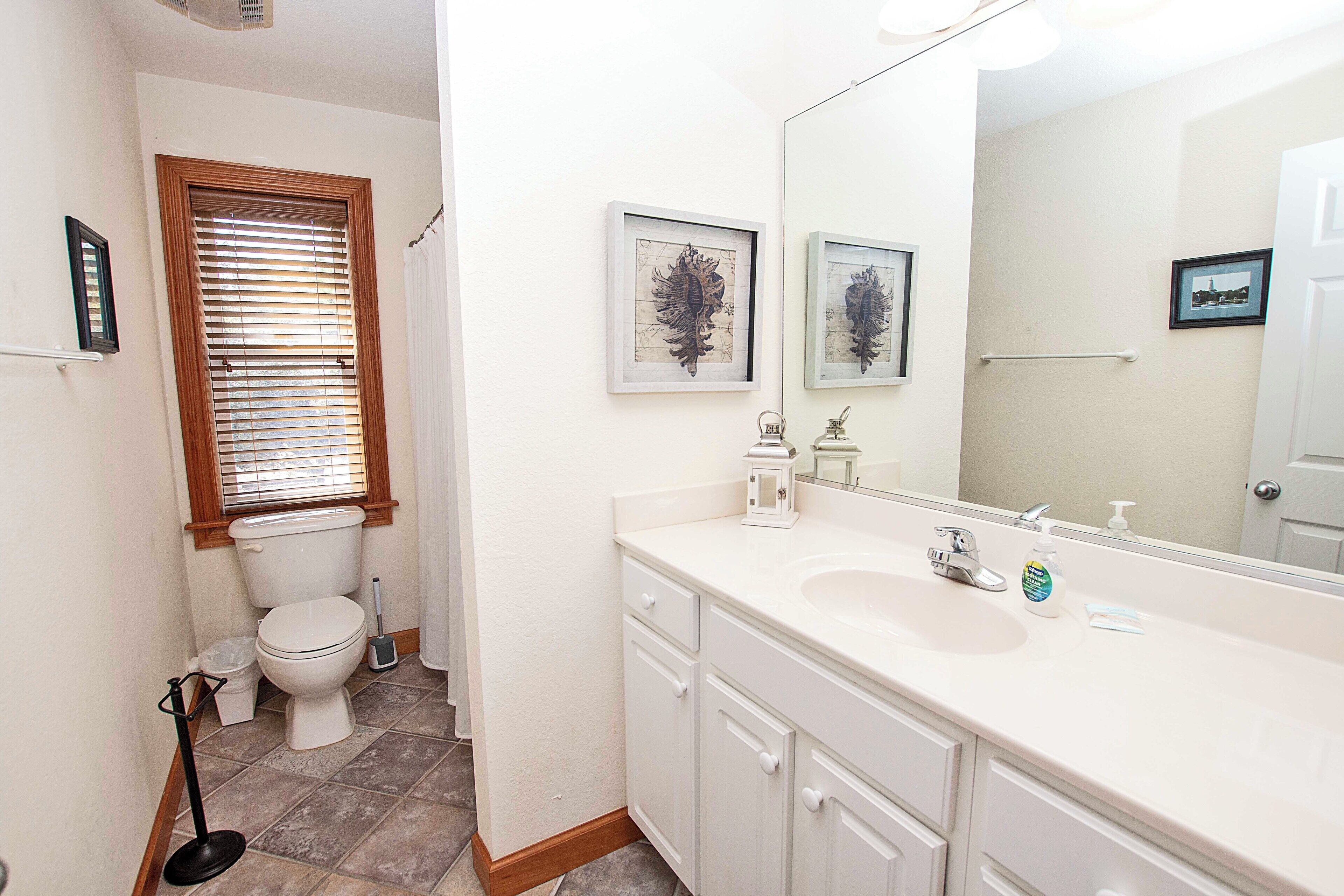 A bathroom with a toilet, sink, and mirror. There are framed pictures on the wall and a window with blinds.