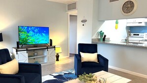 Un salon avec un téléviseur, deux fauteuils, une table basse et, au fond, un coin cuisine.