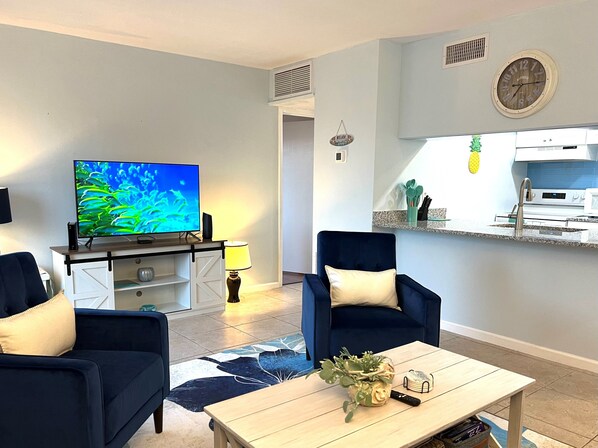 Ein Wohnzimmer mit Fernseher, zwei Sesseln, einem Couchtisch und einer Küche im Hintergrund.