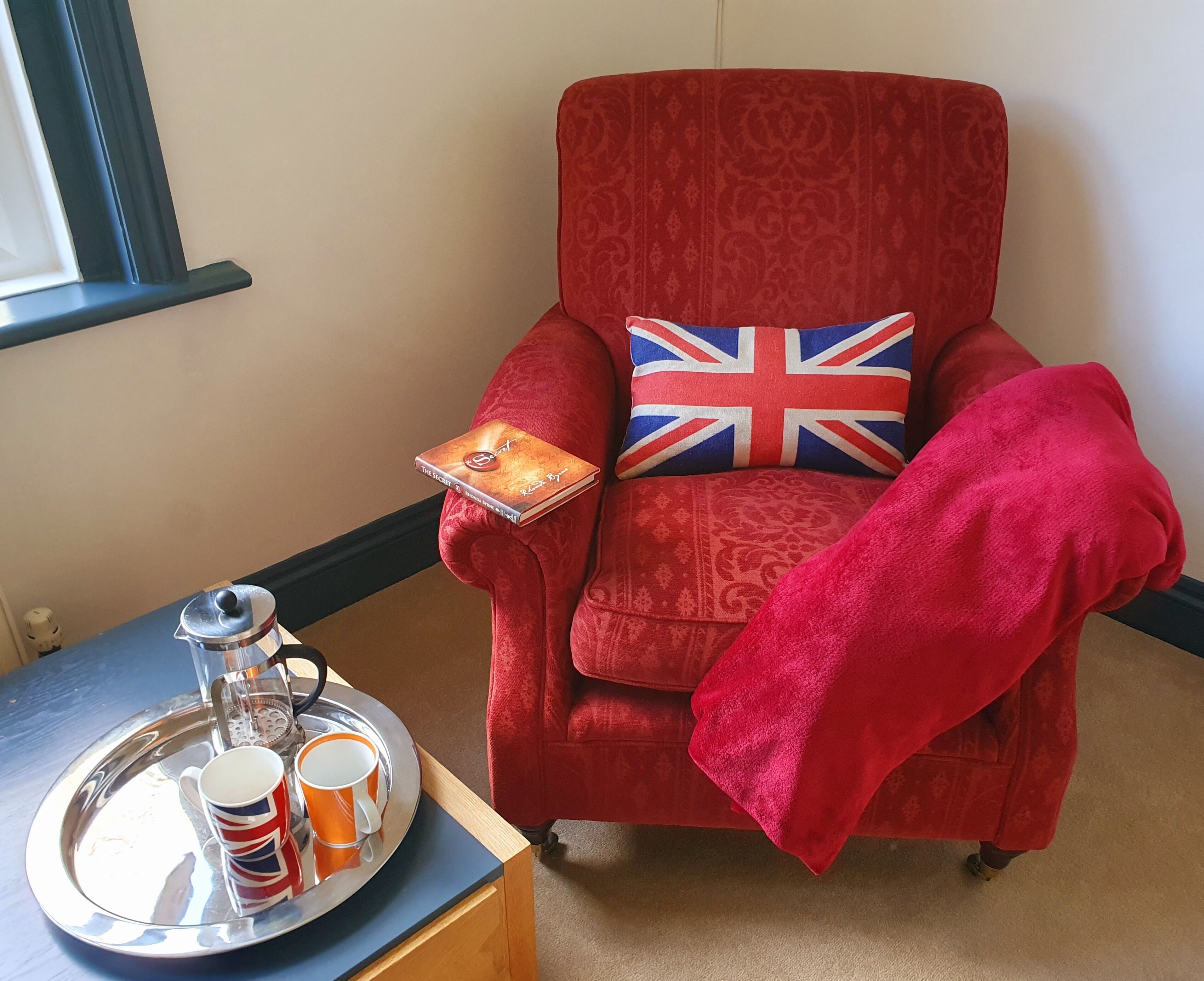 A red armchair with a Union Jack pillow, a tray with mugs and a book, and a window in the background.