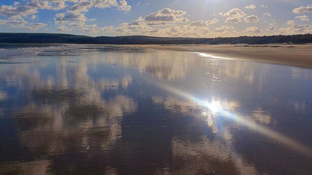 A beach with a calm sea, clear sky, and sunlight reflecting on the wet sand.