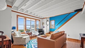 A spacious living room with a high ceiling, large windows, a brown leather sofa, a white armchair, a wooden dining table, and a blue accent wall. - Cozy Nest | Water View Retreat w/ Screened Porch, Private Deck Suite & Pet Friendly Comfort for 6 (Chincoteague Island)
