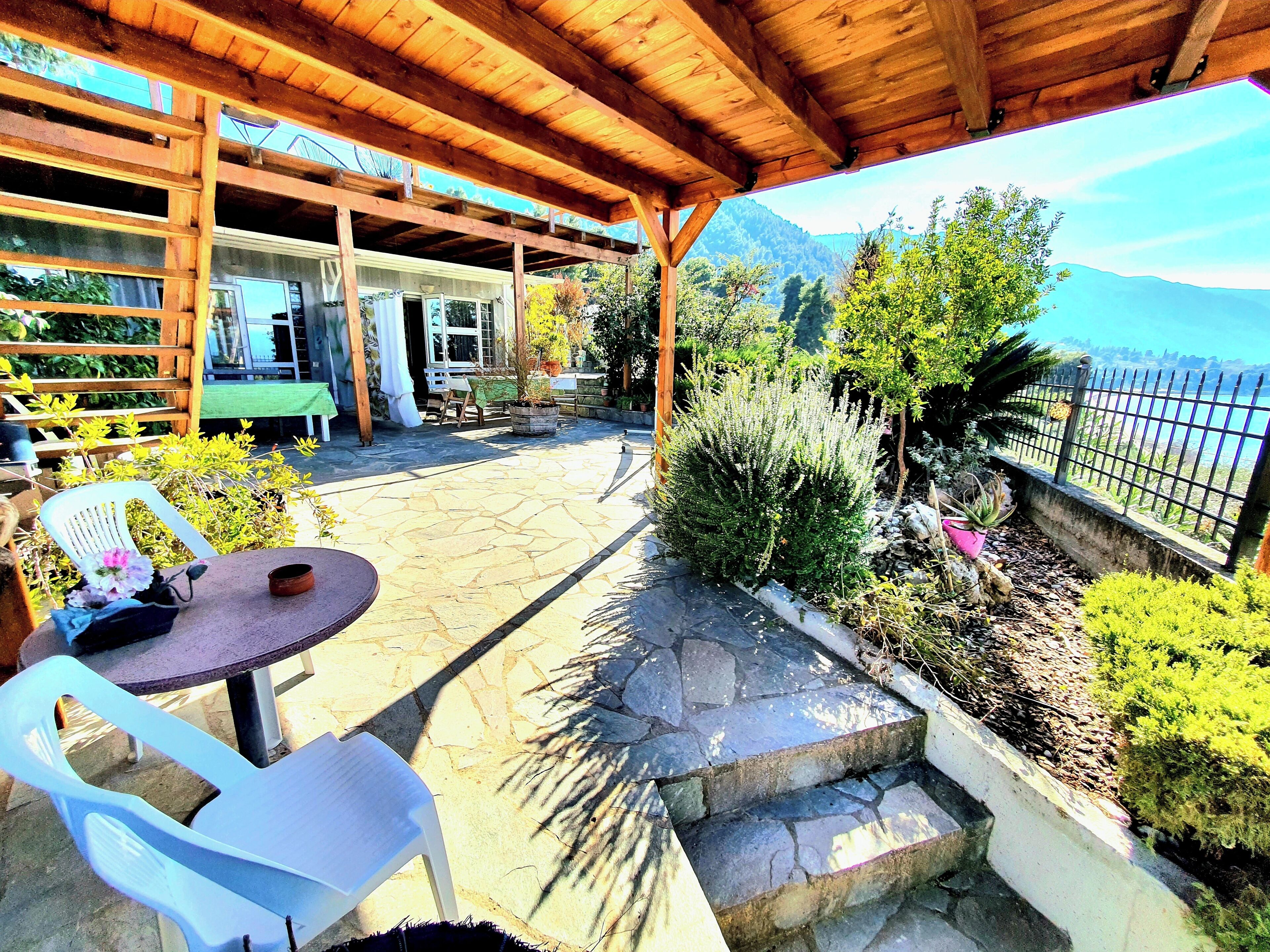 An outdoor patio with stone flooring, wooden pergola, white plastic chairs, and a view of a mountainous landscape.