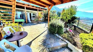 An outdoor patio with stone flooring, wooden pergola, white plastic chairs, and a view of a mountainous landscape.