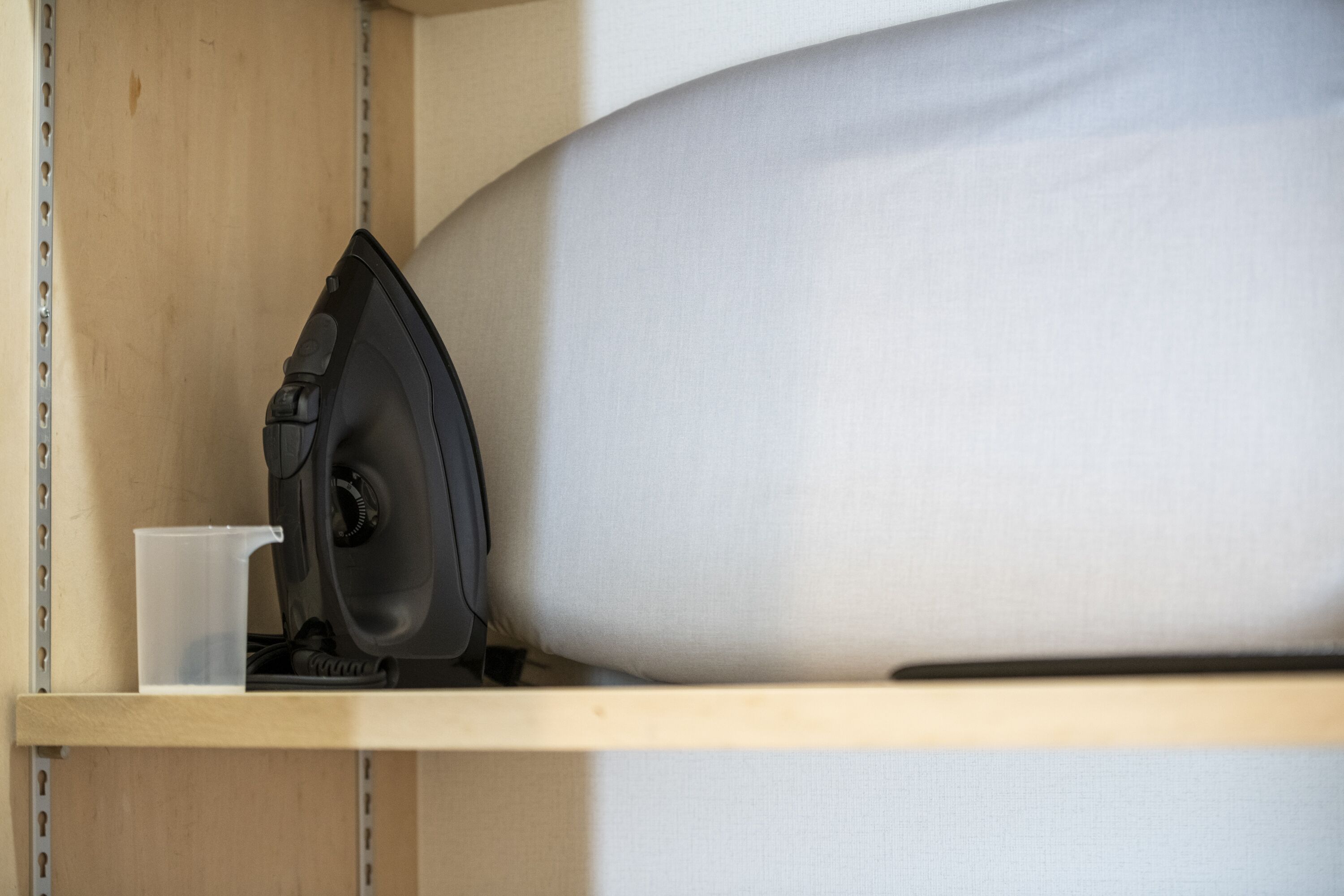 A wooden shelf with a black iron, a clear plastic cup, and a white pillow.