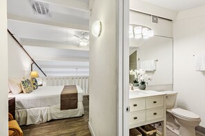 Une chambre à coucher avec un lit, une table de chevet et une commode. Il y a une fenêtre avec des stores et un ventilateur de plafond.