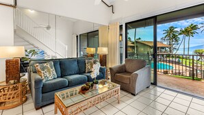 A spacious living room with a blue sofa, a grey armchair, and a glass coffee table. A staircase with white railings leads to a balcony with a pool and palm trees. - Top floor ocean view 2bd condo (Lahaina)