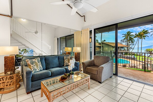 A spacious living room with a blue sofa, a grey armchair, and a glass coffee table. A staircase with white railings leads to a balcony with a pool and palm trees.